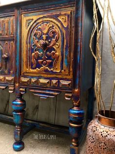 an ornate blue and gold painted cabinet next to a potted plant on the floor