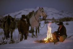 a group of men standing around a fire in the snow with horses behind them on a snowy day