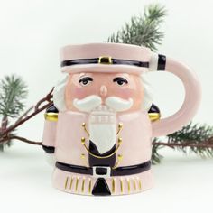 a pink and black coffee mug with a santa clause on it's face, sitting next to a pine branch