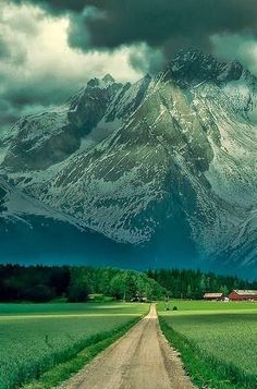 a dirt road in front of a snow covered mountain with green grass on both sides