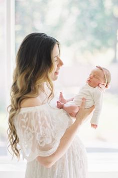 a woman holding a baby in her arms and looking at the camera while wearing a white dress