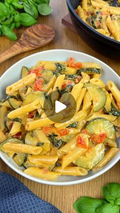 pasta with zucchini and tomatoes in a white bowl on a wooden table next to basil leaves