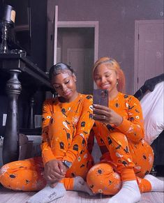 two women in orange pajamas sitting on the floor looking at a cell phone and smiling