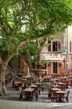 an outdoor dining area with tables, chairs and trees in front of a building on a cobblestone street