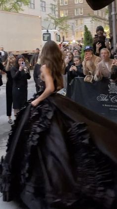 a woman in a black dress walking down the street with people taking pictures behind her