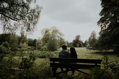 two people sitting on a bench in the middle of a field with trees and grass