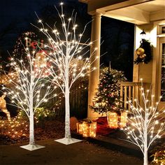 lighted trees in front of a house with christmas decorations on the porch and side walk