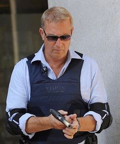 an older man wearing sunglasses and holding a cell phone in his hand while standing next to a building