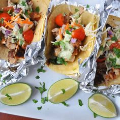 three tacos with meat, vegetables and cheese on tin foil next to lime wedges