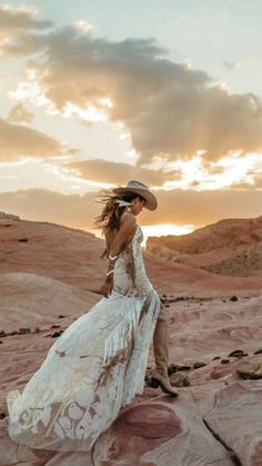 a woman in a white dress and hat standing on rocks with the sun setting behind her