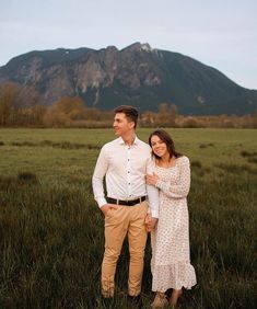 a man and woman standing next to each other in a field with mountains in the background