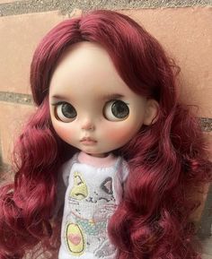 a close up of a doll with long red hair and big eyes, sitting against a brick wall