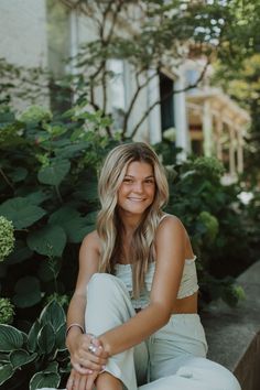 a woman sitting on the ground in front of some bushes and plants smiling at the camera