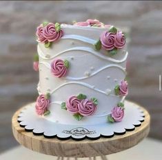 a white cake with pink roses on it sitting on top of a wooden stand next to a brick wall