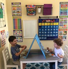 two young boys sitting at a table playing with toys in their playroom, while calendars hang on the wall behind them