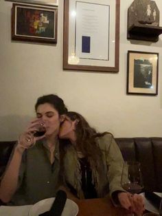 two women sitting at a table with wine glasses in front of their faces, one woman is kissing the other