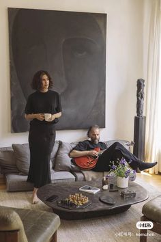 a woman standing next to a man sitting on a couch in front of a painting