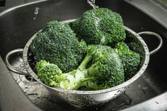 broccoli is being washed in a metal bowl on the kitchen sink with water splashing over it