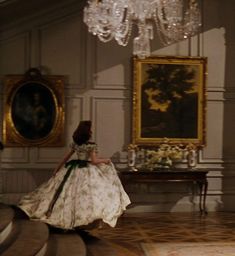 a woman in a white dress walking through a room with chandelier and paintings on the wall