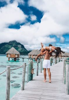 a man and woman are standing on a dock in front of some water with thatched huts