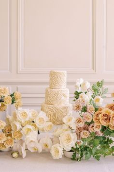 a wedding cake surrounded by flowers and greenery on a white tablecloth with wall in the background