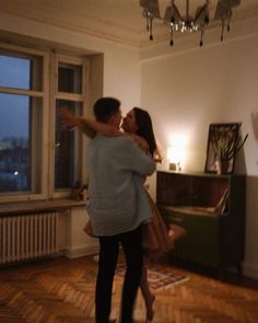 a man and woman dancing in a room with wooden floors, chandelier and windows