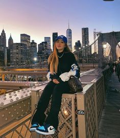 a woman sitting on top of a metal fence next to a bridge with the city skyline in the background