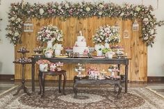 a table topped with lots of cakes and flowers next to a wall covered in greenery