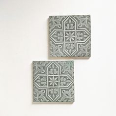 two square tiles with designs on them sitting side by side against a white wall, one in grey and the other in light green