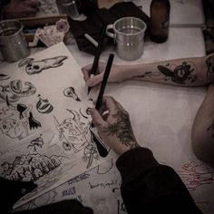 two people sitting at a table with tattoos on their arms and legs, one holding a pen