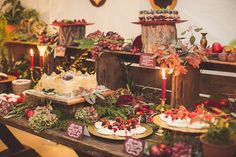 a table topped with lots of cakes and desserts covered in greenery next to candles