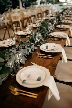 the table is set with white plates and place settings, greenery on each plate