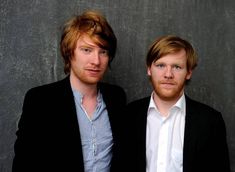 two men standing next to each other in front of a gray wall and one is looking at the camera