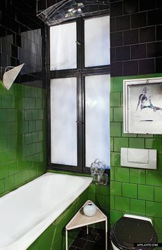 a bathroom with green tiles and white tub next to a black trash can in front of a window