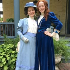 two women dressed in period clothing standing next to each other on a brick walkway near bushes