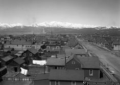 an old black and white photo of a town with snow capped mountains in the background