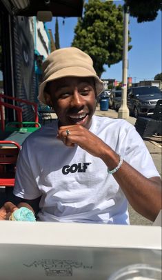 a man sitting in front of a white car eating something out of his hand while wearing a hat