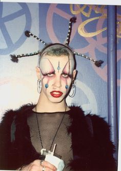 a woman with clown makeup and piercings holding a drink in her hand while standing next to a wall