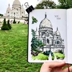 a hand holding up an open book in front of a grassy area with trees and buildings