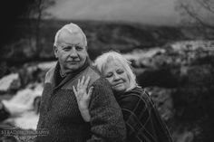 an older man and woman standing next to each other near a river with mountains in the background