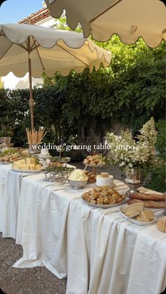 an outdoor buffet table with cheeses and bread on plates under umbrellas in the shade