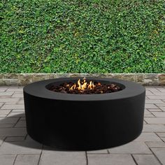 an outdoor fire pit sitting on top of a brick floor next to a green hedge