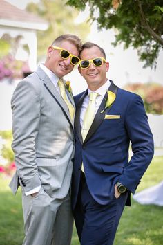 two men in suits and yellow sunglasses posing for the camera on some grass with trees in the background