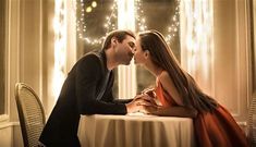 a man and woman kissing each other at a table with lights on the wall behind them