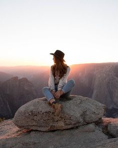 a woman sitting on top of a large rock next to a mountain at sunset with the sun behind her