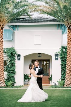 a bride and groom standing in front of a large white house with palm trees on the lawn