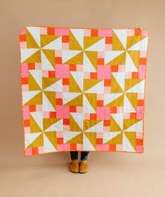 a person holding up a yellow and pink quilt in front of a tan background with white triangles on it