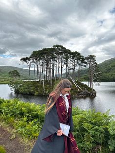 a woman dressed in medieval clothing is walking by the water with trees and bushes behind her