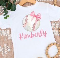 "Get ready to score big style points this fall with our charming and personalized girls' baseball shirt! Your little football fan will look adorable and sporty with a baseball featuring a cute bow and her own custom name printed underneath. Customization: Make this shirt uniquely hers by adding her name! Simply leave the desired name in the \"Note to Seller\" section during checkout, and we'll create a one-of-a-kind shirt that she'll love to wear all season long. Quality and Comfort: Our shirts are crafted with the utmost care from soft and breathable cotton fabric, ensuring your little one's comfort during all her baseball-related activities. Whether she's cheering on her favorite team or playing a friendly game with friends, she'll feel at ease in this cozy and stylish shirt. Design Deta Personalized White Sporty T-shirt, Sporty Personalized T-shirt For Game Day, Personalized Sporty T-shirt For Game Day, White Personalized Sporty Top, Sporty Personalized T-shirt For Sports, Personalized Sporty T-shirt For Sports, Sporty Personalized Short Sleeve Top, Sporty Personalized White Top, Softball Bow