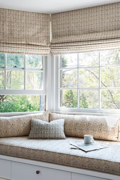 a window seat with pillows on it in front of two windows, one has a coffee cup and the other is a book